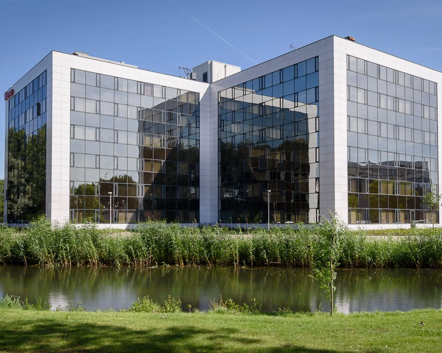 Modern kantoorgebouw met spiegelende glazen gevels aan het water, onder een blauwe lucht.