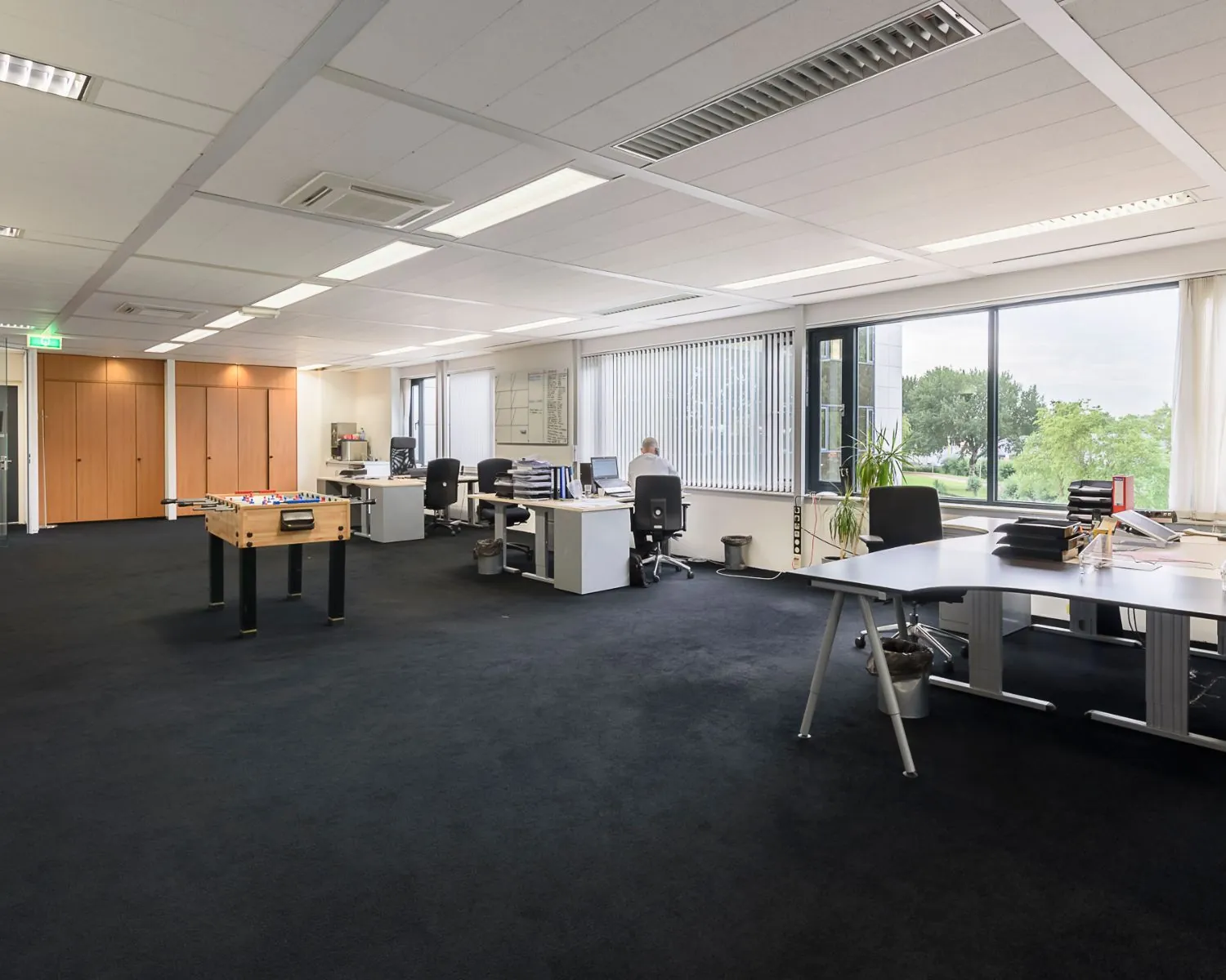 Ruim en licht modern kantoor met werkplekken, grote ramen en een tafelvoetbaltafel in het midden.