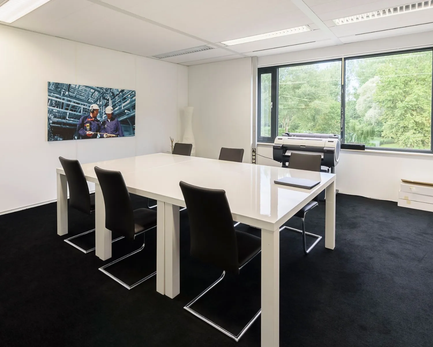 Moderne vergaderruimte met wit tafelblad, zwarte stoelen, groot raam en een foto van twee industriële arbeiders aan de muur.