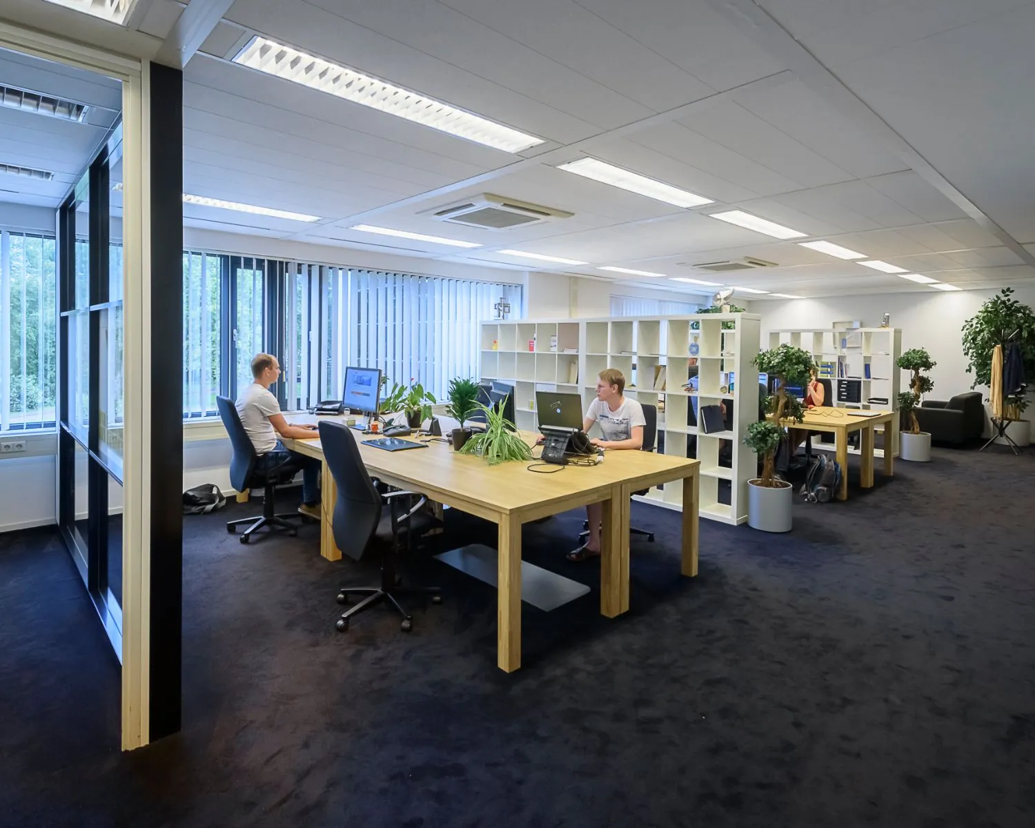 Moderne, open kantoorruimte met houten bureaus, computers, planten en werkende mensen.