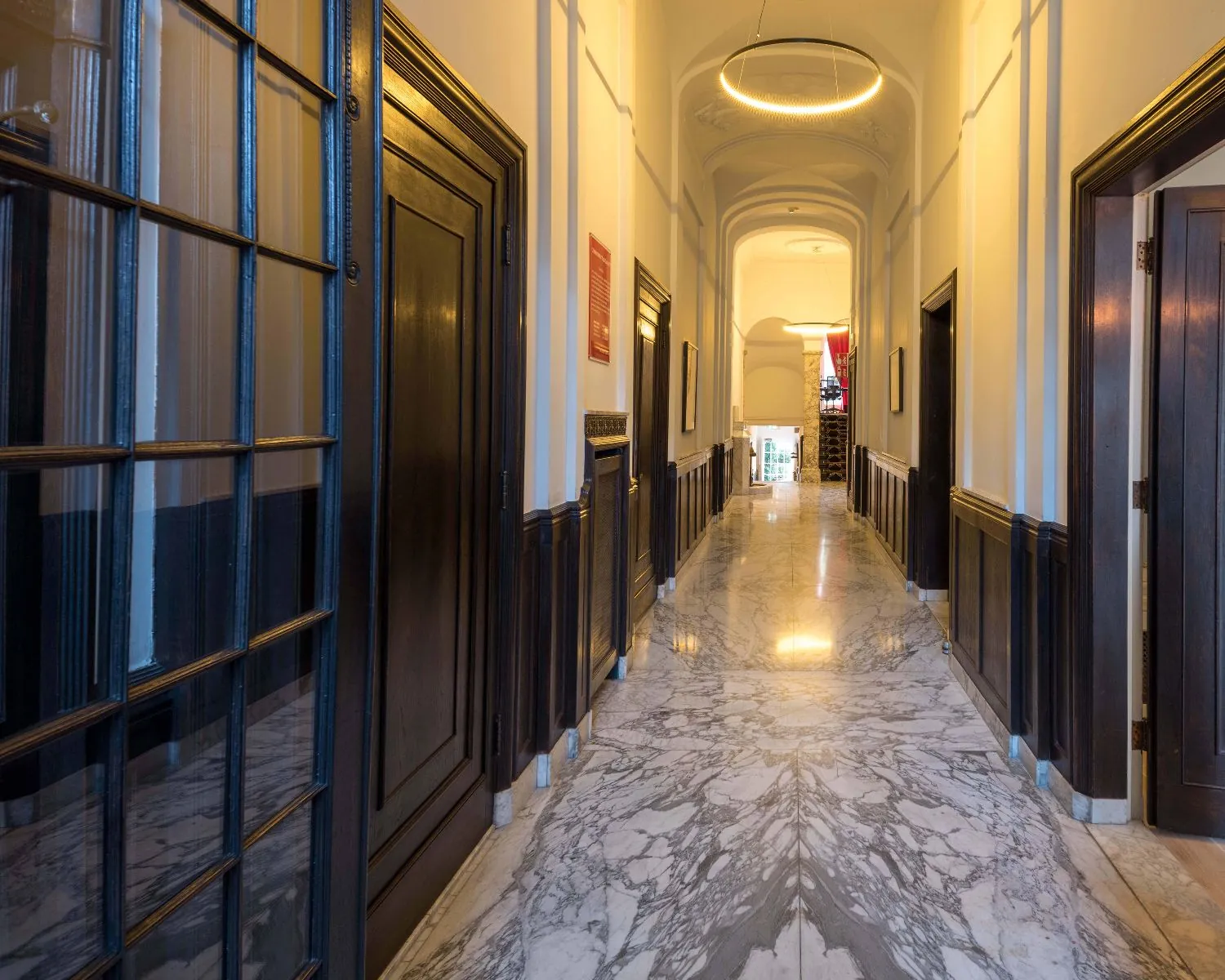 Elegante marmeren gang met hoge plafonds, houten deuren en een moderne ronde hanglamp.