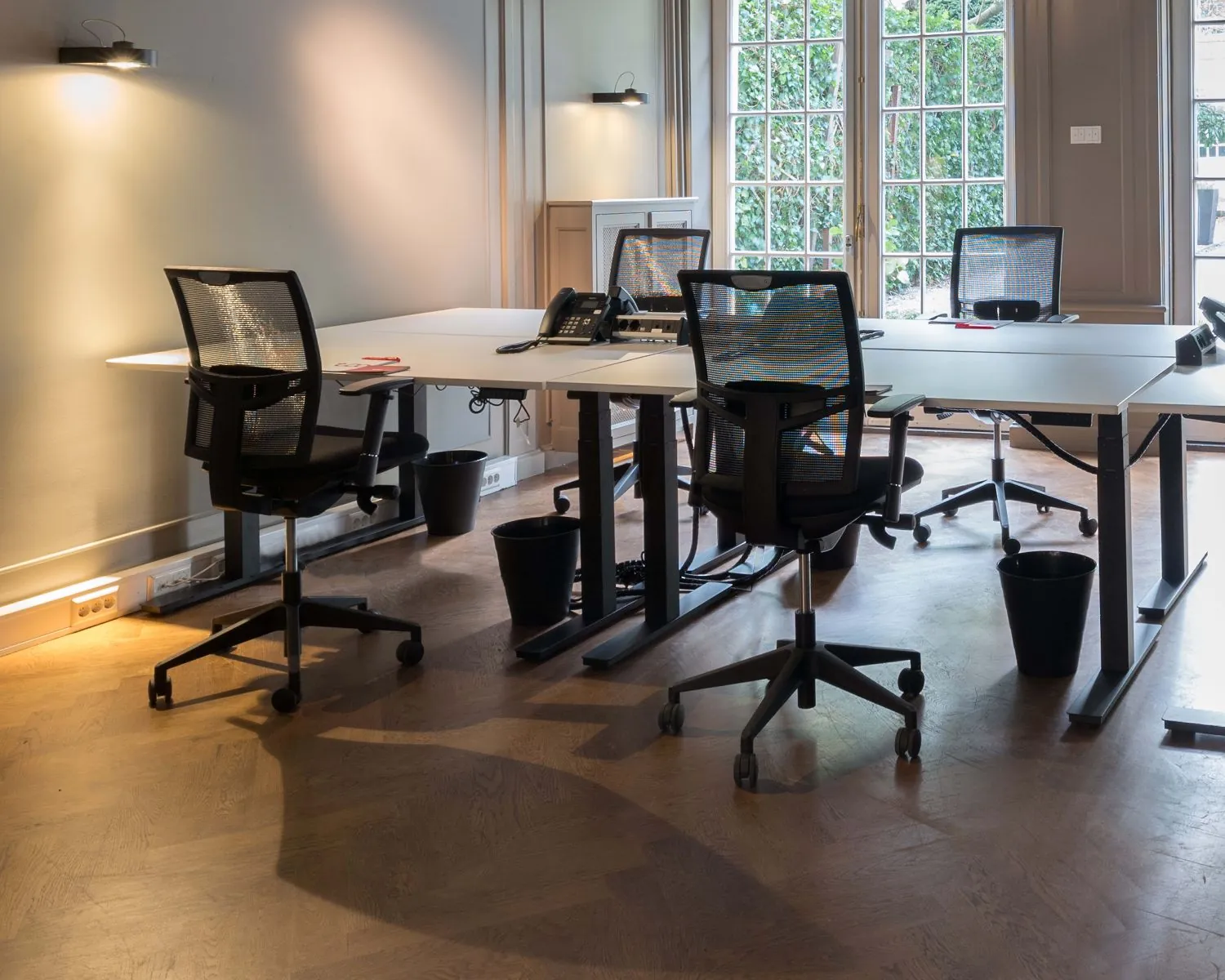 Moderne, lichte kantoorruimte met bureaus, bureaustoelen en telefoons, gelegen bij een raam met uitzicht op groen.