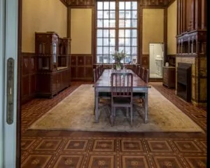 Historische eetkamer met houten lambrisering, lange eettafel, hoge ramen en rijk versierde vloer en schouw.