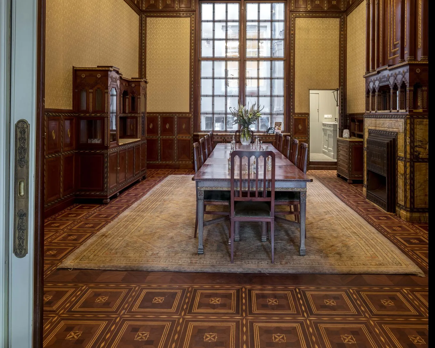 Historische eetkamer met houten lambrisering, lange eettafel, hoge ramen en rijk versierde vloer en schouw.