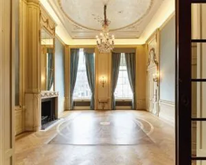 Elegante, klassieke balzaal met hoge plafonds, kroonluchters, open haard en grote ramen met blauwe gordijnen.