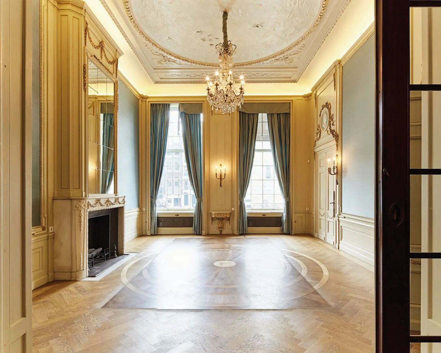 Elegante, klassieke balzaal met hoge plafonds, kroonluchters, open haard en grote ramen met blauwe gordijnen.