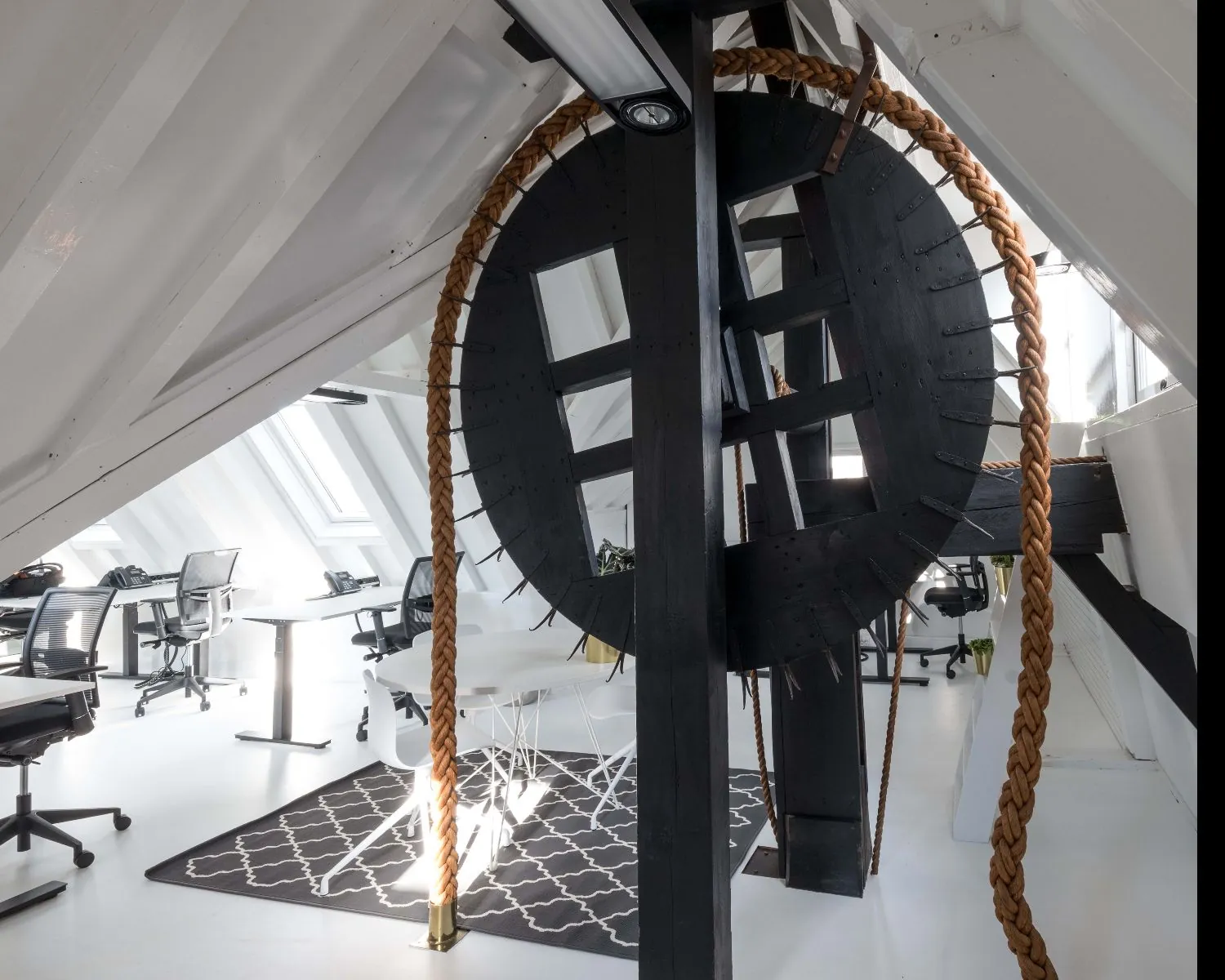 Modern kantoor in een witgeschilderde zolderruimte met een groot zwart houten hijswiel en touw als opvallend industrieel element.
