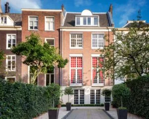 Achteraanzicht van een klassiek herenhuis met rode gordijnen en een verzorgde tuin met bomen en planten in potten.