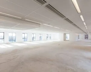 Lichte, lege kantoorruimte met grote ramen en systeemplafond.