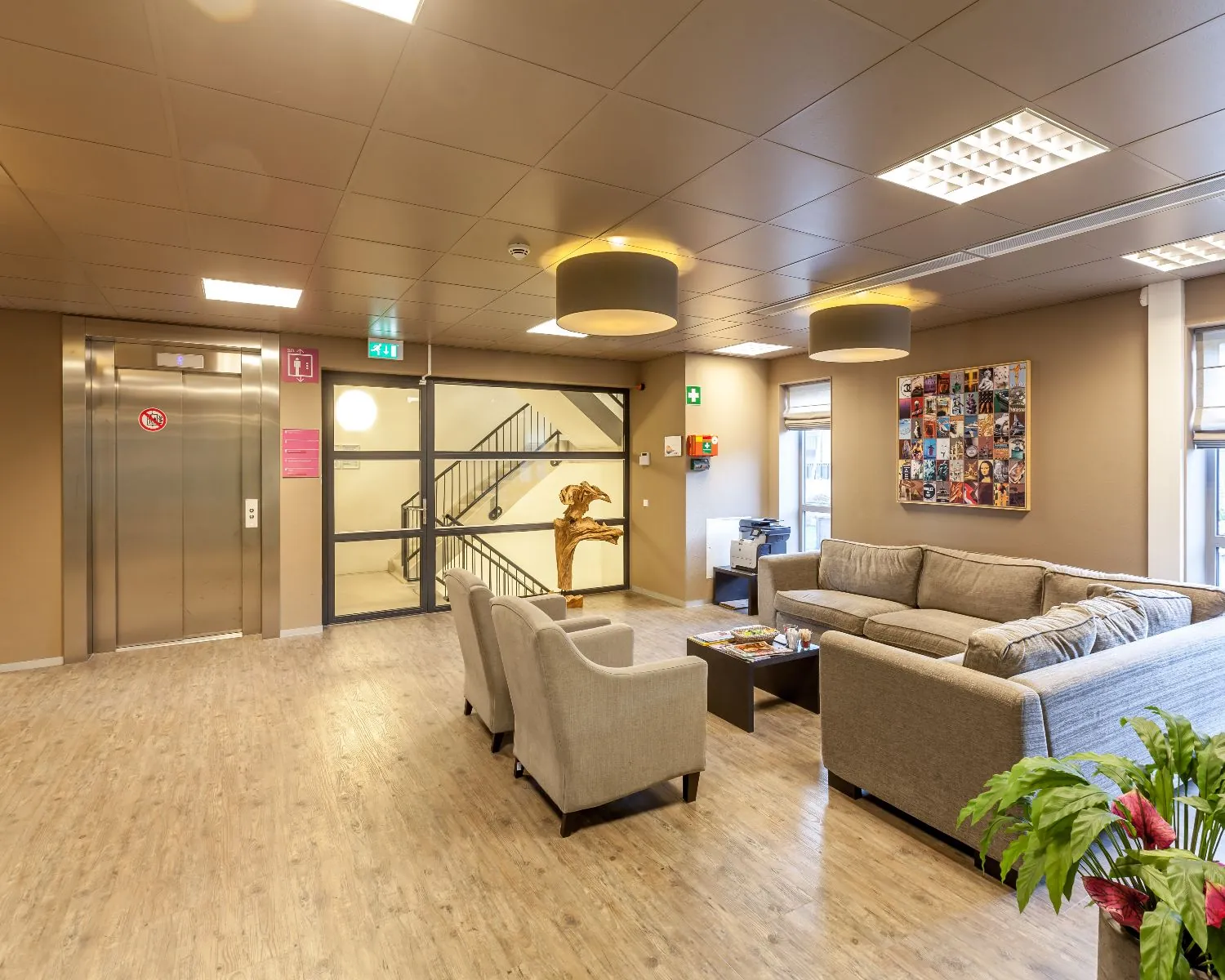 Wacht- of ontvangstruimte met zitbanken, fauteuils, een lift en een kunstwerk aan de muur.