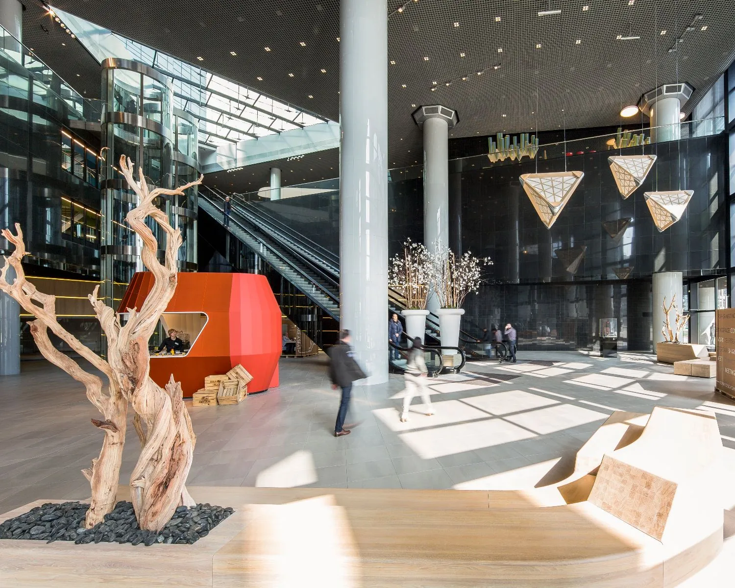 Moderne, lichte lobby met kunstobjecten, een rood kioskachtig bouwwerk en roltrappen naar boven.