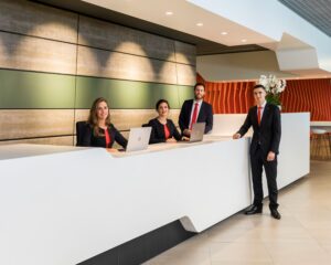Vier medewerkers in zakelijke kleding staan en zitten achter een moderne receptiebalie in een strak vormgegeven kantoorruimte.