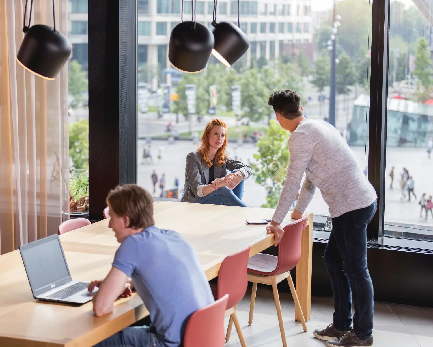 Drie jonge mensen overleggen informeel in een moderne kantoorruimte met uitzicht op een stadsplein.