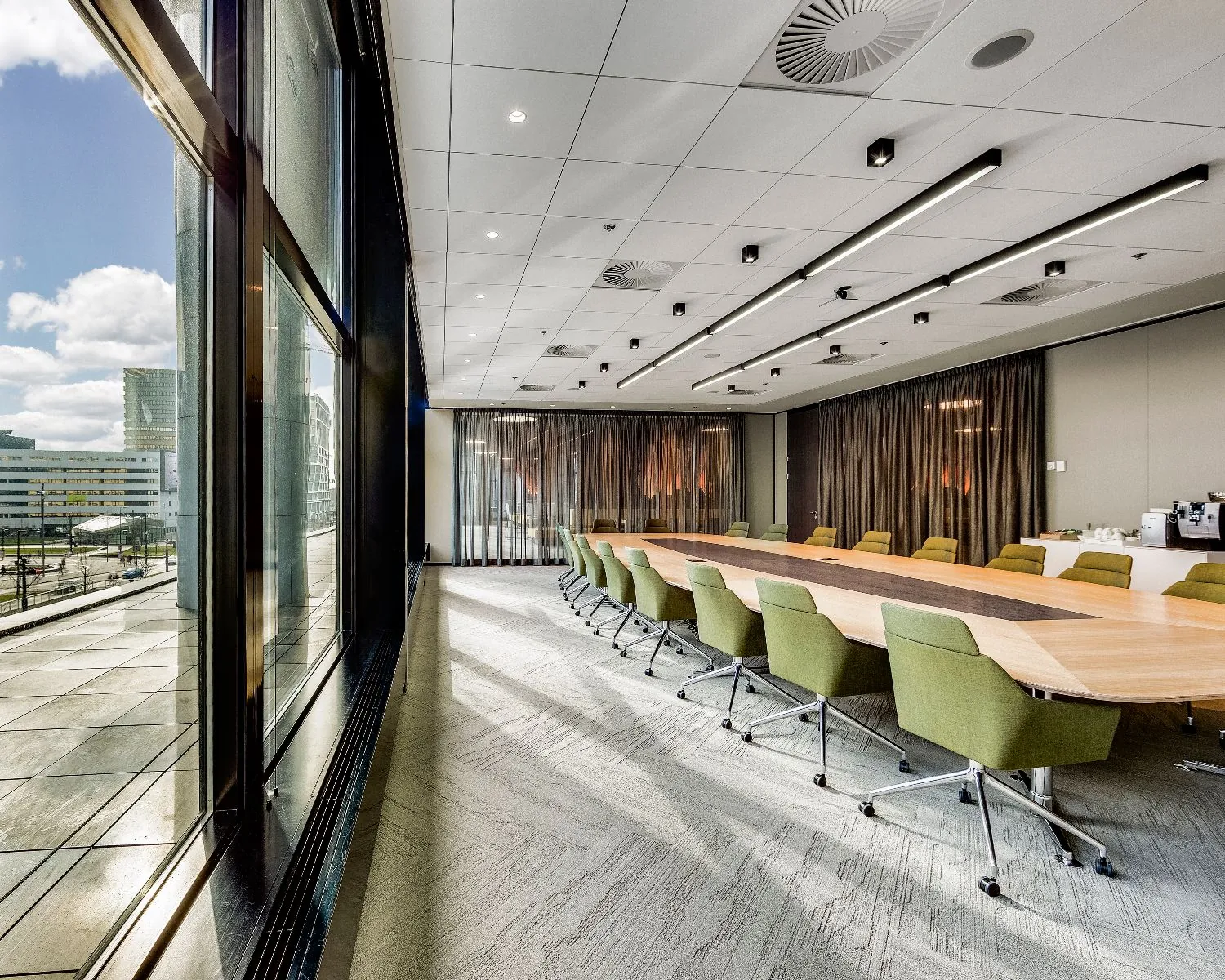 Luxe vergaderruimte met lange tafel, groene stoelen en grote ramen met uitzicht op moderne kantoorpanden.