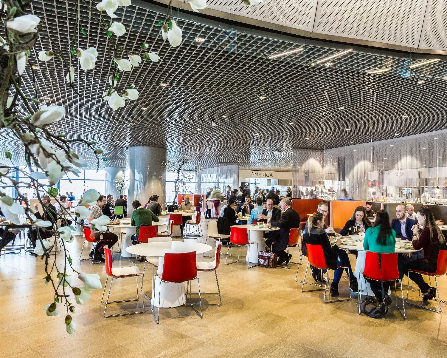 Moderne, drukbezochte bedrijfskantine met mensen die aan ronde tafels zitten en eten.