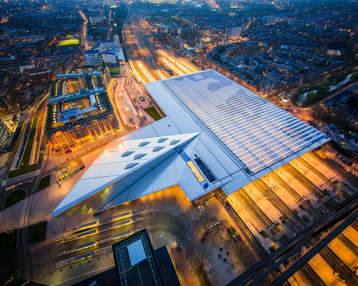 Luchtfoto van station Rotterdam Centraal in de avond met verlichte perrons en omliggende stadsbebouwing.