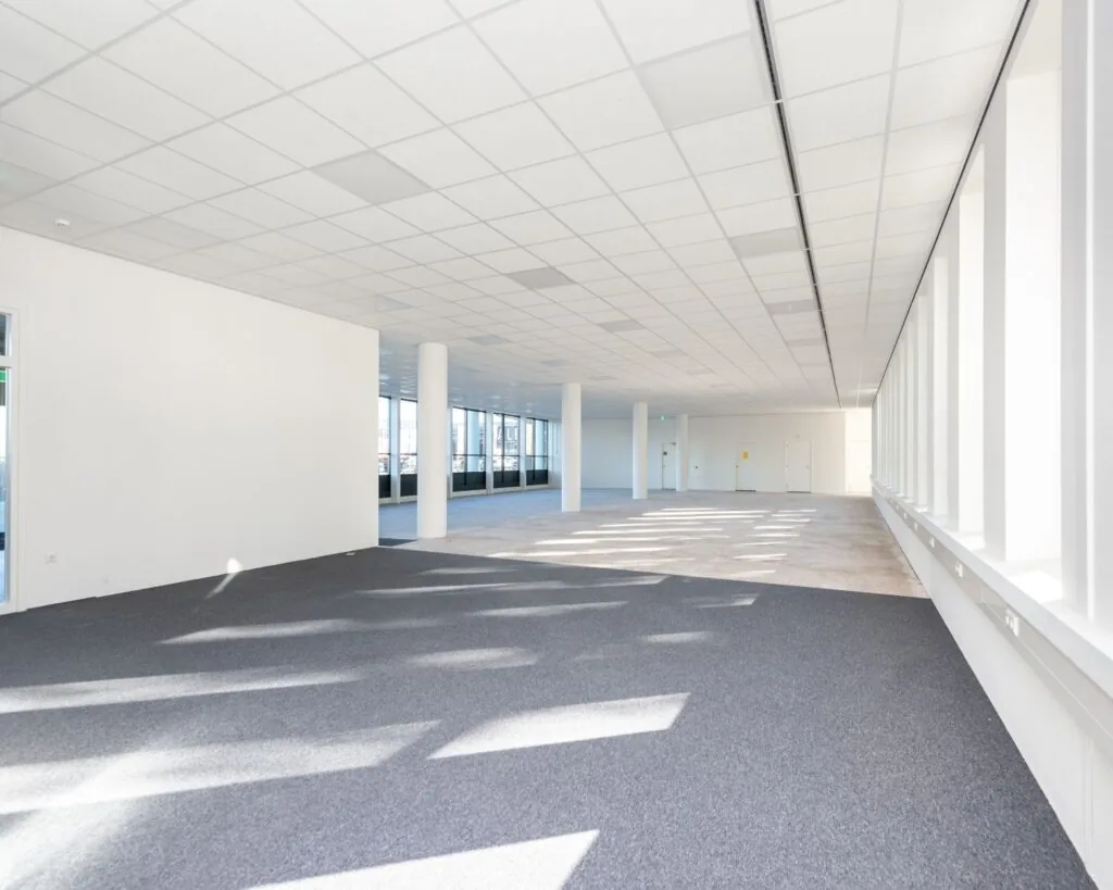 Lichte, lege kantoorruimte met systeemplafond, grote ramen en deels grijze vloerbedekking.
