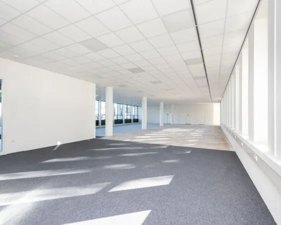 Lichte, lege kantoorruimte met systeemplafond, grote ramen en deels grijze vloerbedekking.