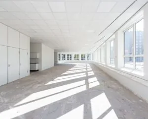 Lichte, lege kantoorruimte met grote ramen en een onafgewerkte betonnen vloer.