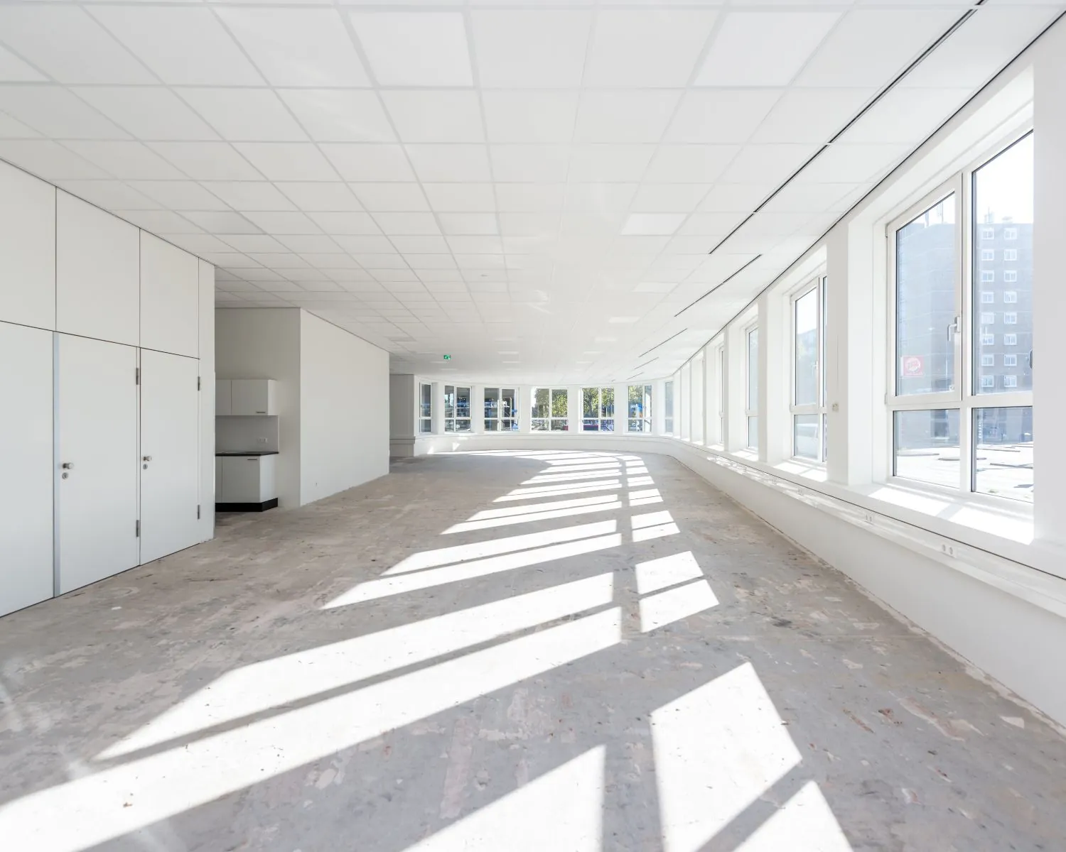 Lichte, lege kantoorruimte met grote ramen en een onafgewerkte betonnen vloer.