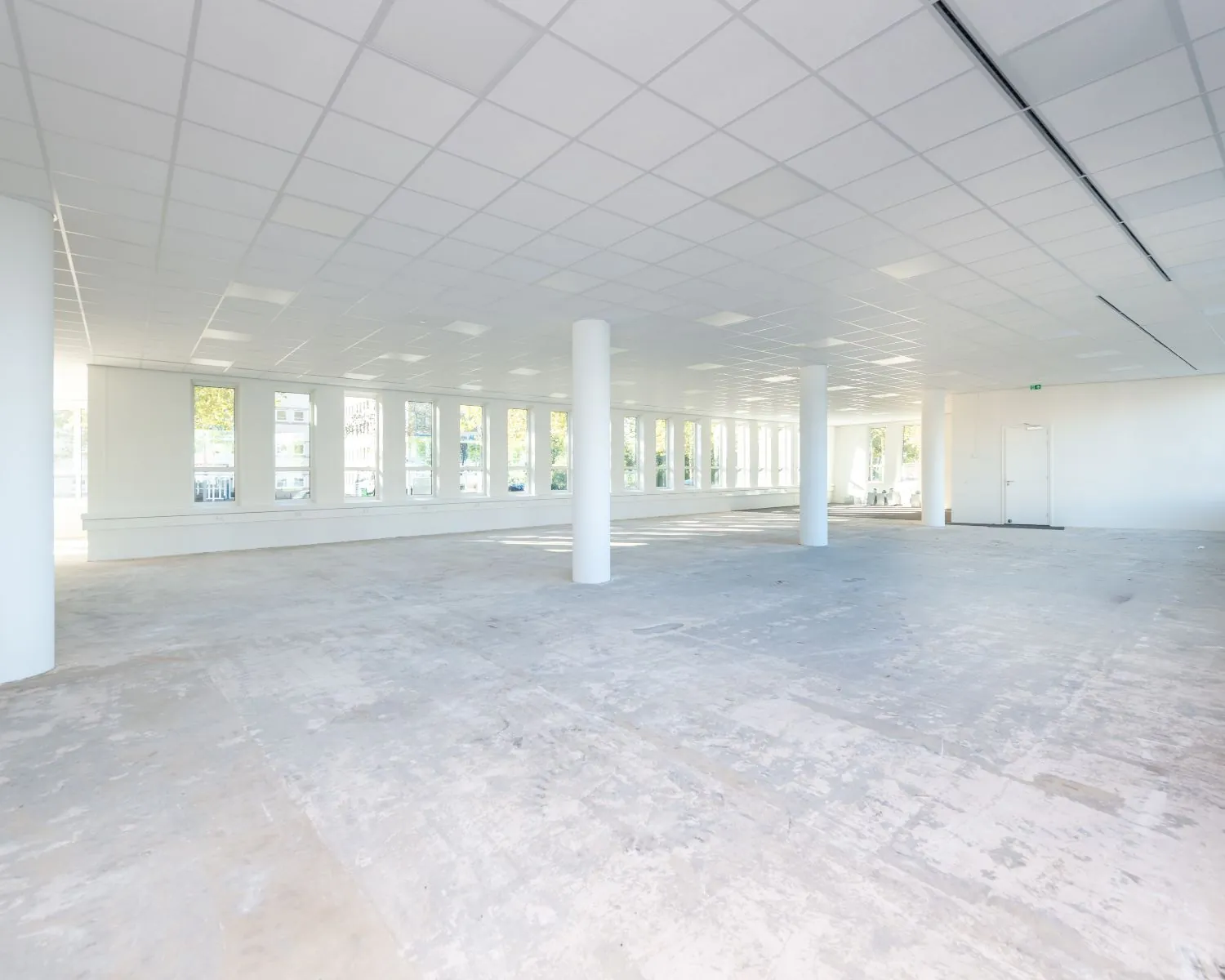 Lichte, lege kantoorruimte met grote ramen en zichtbare betonnen vloer.