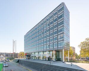 Modern kantoorgebouw met glazen gevel en trappen aan de voorzijde, gelegen in een stedelijke omgeving.