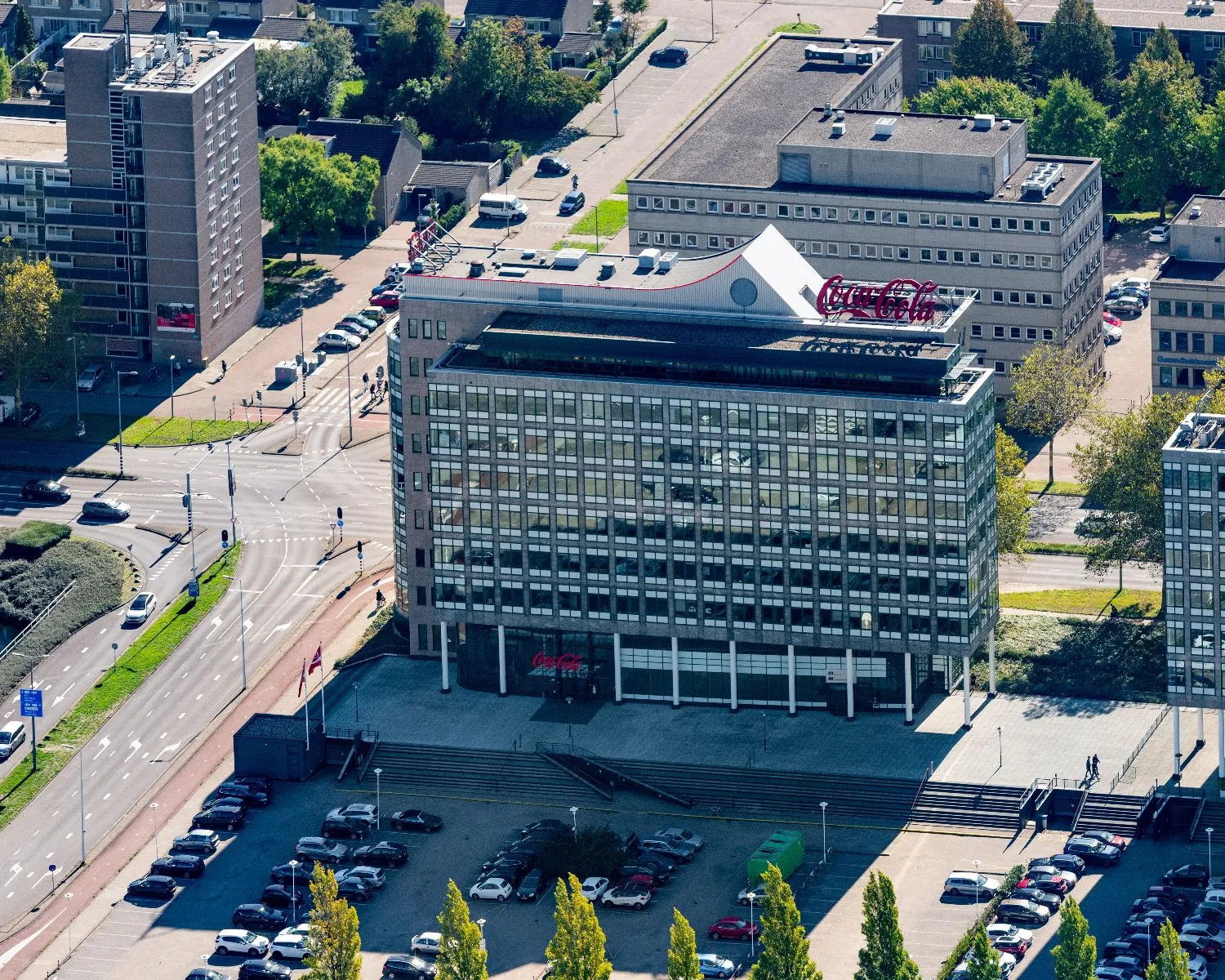 Luchtfoto van een kantoorgebouw met Coca-Cola-logo op het dak, gelegen aan een druk kruispunt met omliggende parkeerplaatsen en bebouwing.