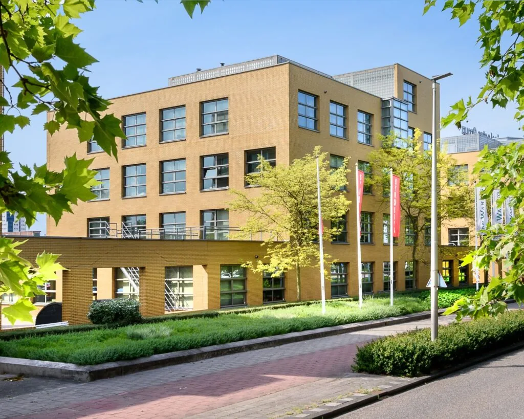 Modern schoolgebouw met gele bakstenen gevel en kleurrijke vlaggen, omgeven door groen.