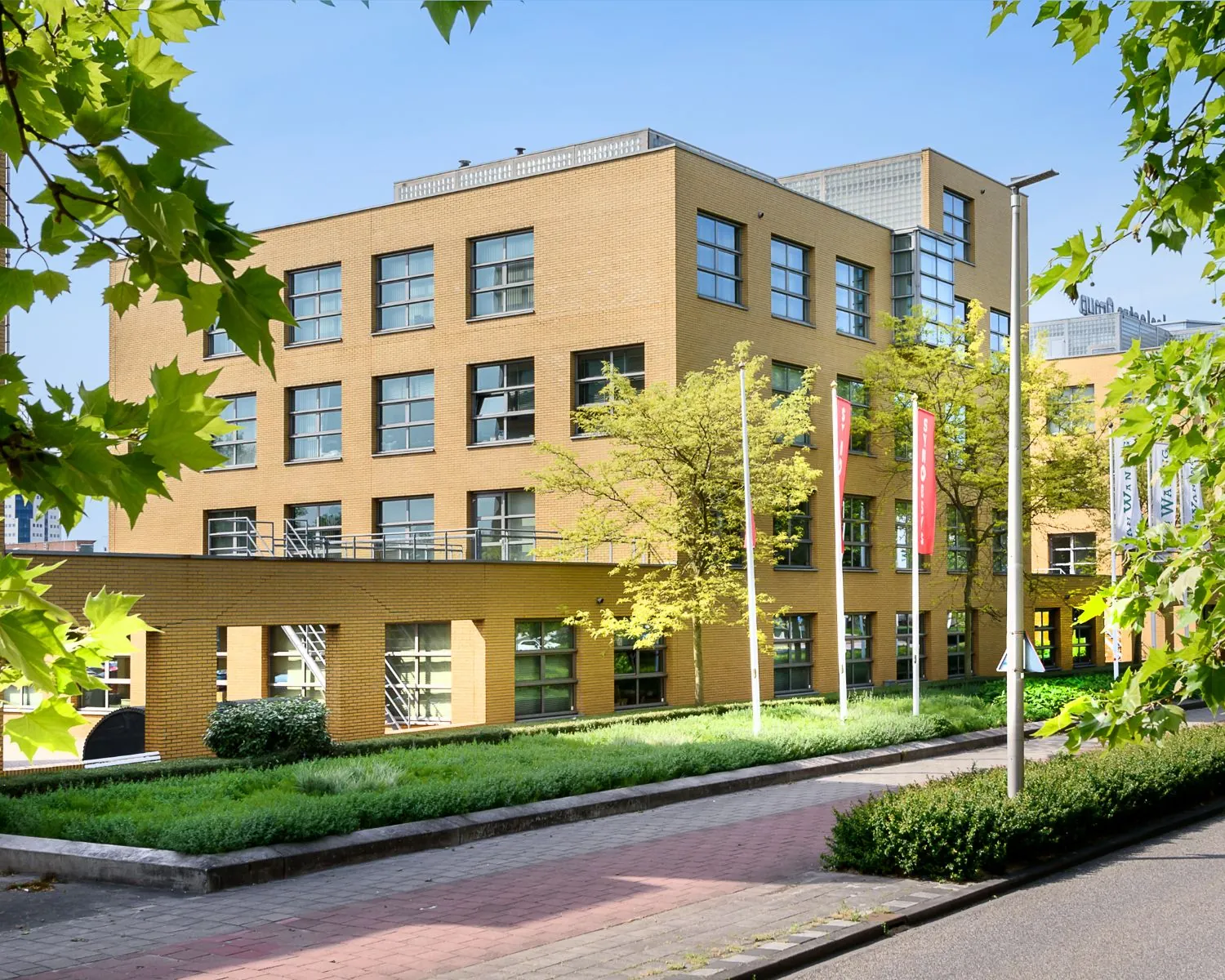 Modern schoolgebouw met gele bakstenen gevel en kleurrijke vlaggen, omgeven door groen.
