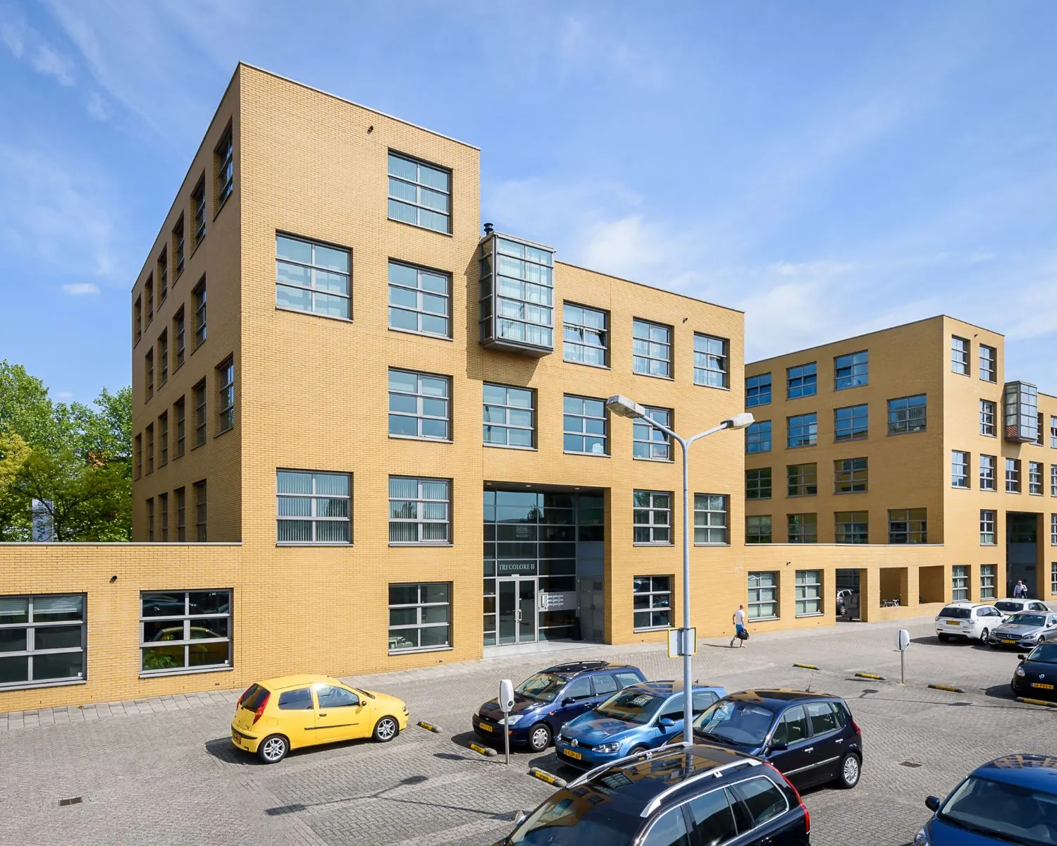 Modern kantoorgebouw met een gele bakstenen gevel en meerdere geparkeerde auto’s op de voorgrond.