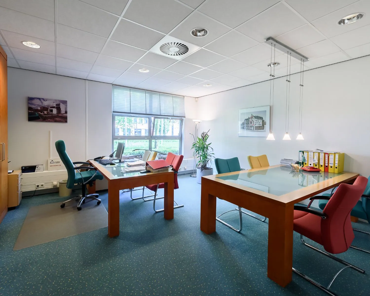 Modern kantoorinterieur met twee bureaus, kleurrijke stoelen en grote ramen die daglicht binnenlaten.