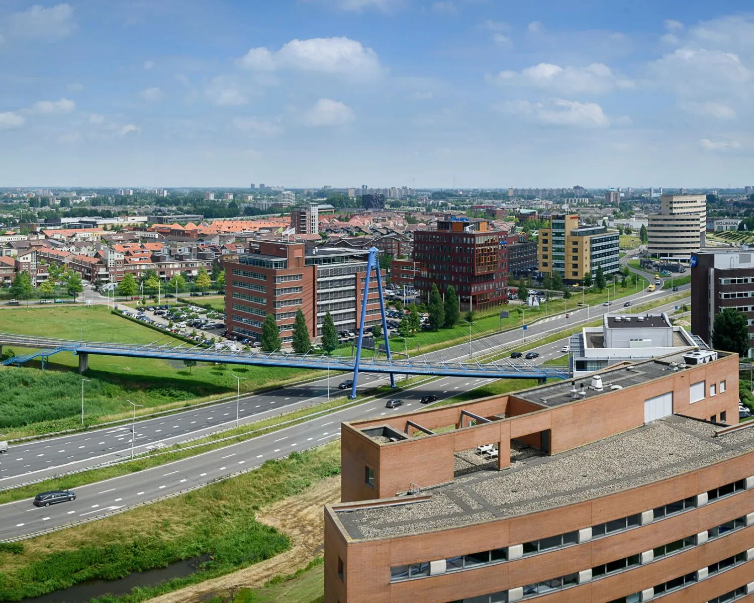 Luchtfoto van een modern zakendistrict met kantoren, woonwijken en een opvallende blauwe voetgangersbrug over een snelweg.