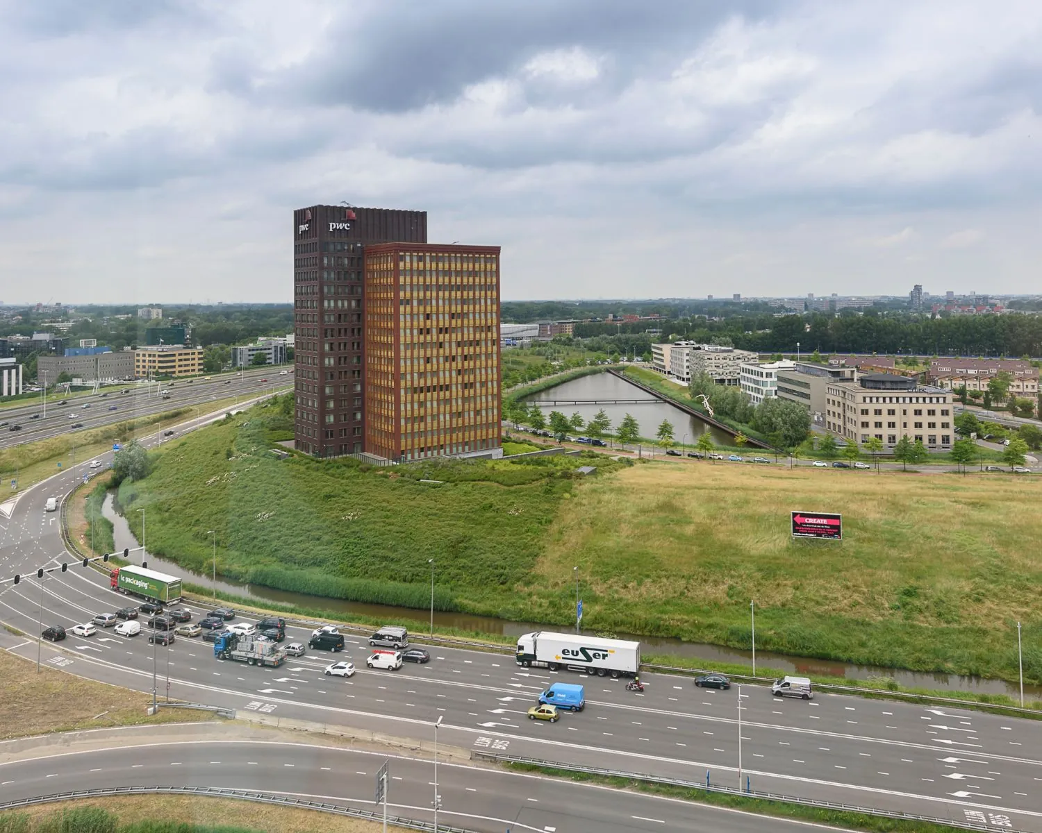 Luchtfoto van een kantoorgebouw van PwC naast een druk kruispunt en snelweg in een groen stedelijk gebied.