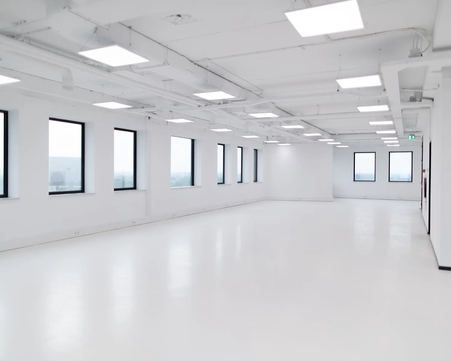 Lichte, lege kantoorruimte met witte vloeren, witte muren en grote ramen.
