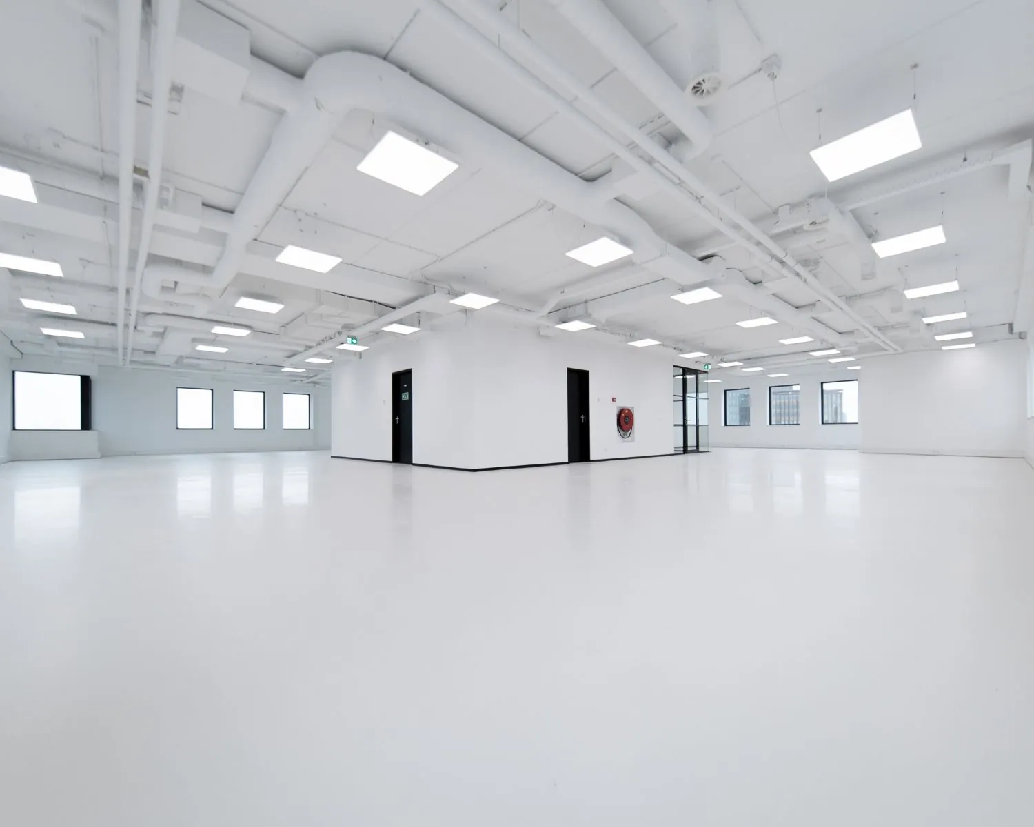 Ruime, lege witte kantoorruimte met zwarte deuren, ramen en zichtbare plafonduitrusting.
