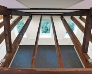 Zolderruimte met zicht op de vloer tussen houten balken en twee ramen met uitzicht op groene bomen.