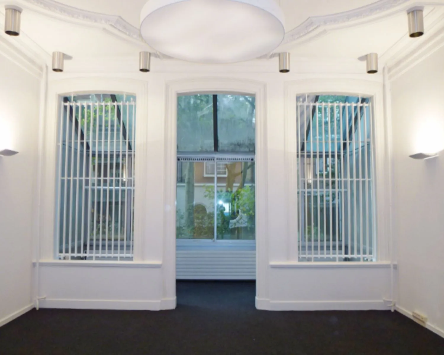 Witte, lege kamer met grote ramen en verticale jaloezieën die uitkijken op een groene binnenplaats.