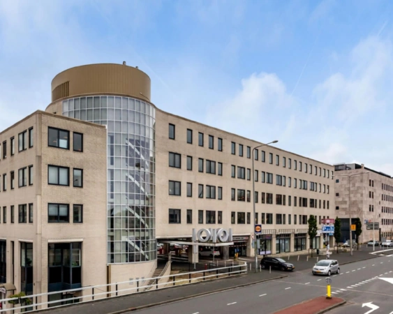 Kantoorgebouw met ronde glazen trappentoren en parkeergarage-ingang aan een drukke stadsweg.