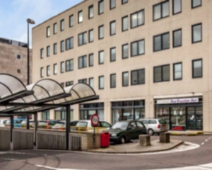 Ingang van een kantoorgebouw met geparkeerde auto's en een overdekte looproute ervoor.