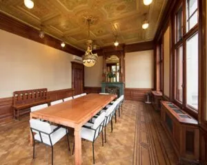 Vergaderruimte met een lange houten tafel, witte stoelen en een rijk gedecoreerd plafond in klassieke stijl.