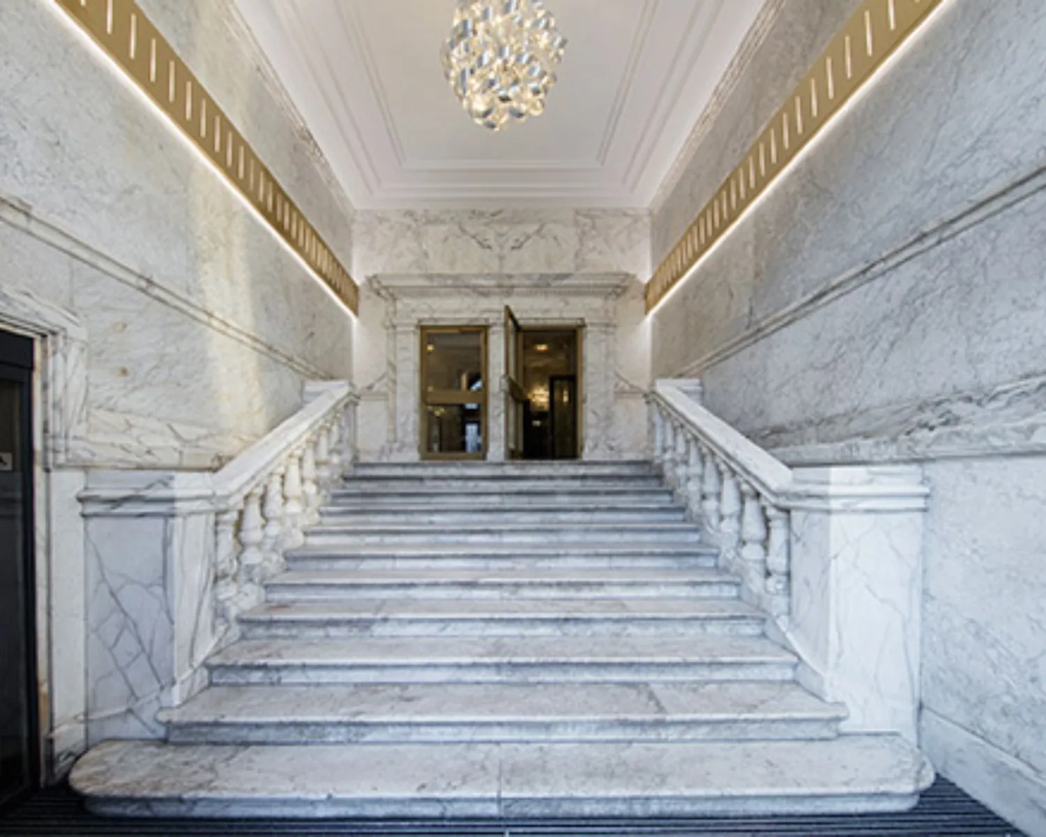 Marmeren trap met balustrades leidt naar een dubbele deur in een luxe, klassiek interieur met een glazen kroonluchter.