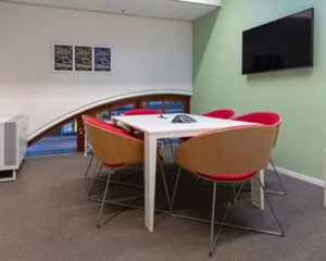 Vergaderruimte met zes moderne stoelen rond een witte tafel, een muurtelevisie en een raam met ronde boog.