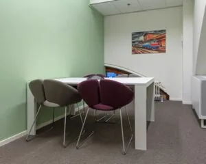 Kleine vergaderruimte met een witte tafel, vier moderne stoelen en een schilderij van een trein aan de muur.