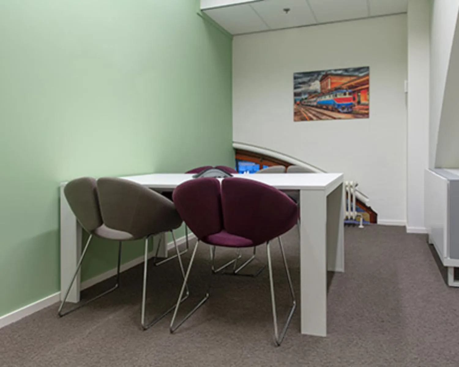 Kleine vergaderruimte met een witte tafel, vier moderne stoelen en een schilderij van een trein aan de muur.