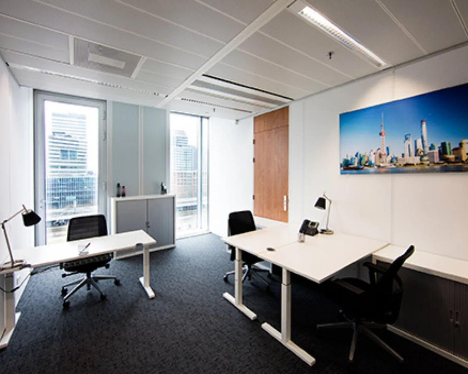 Modern ingericht kantoor met drie bureaus, zwarte bureaustoelen en een grote skylinefoto aan de muur.