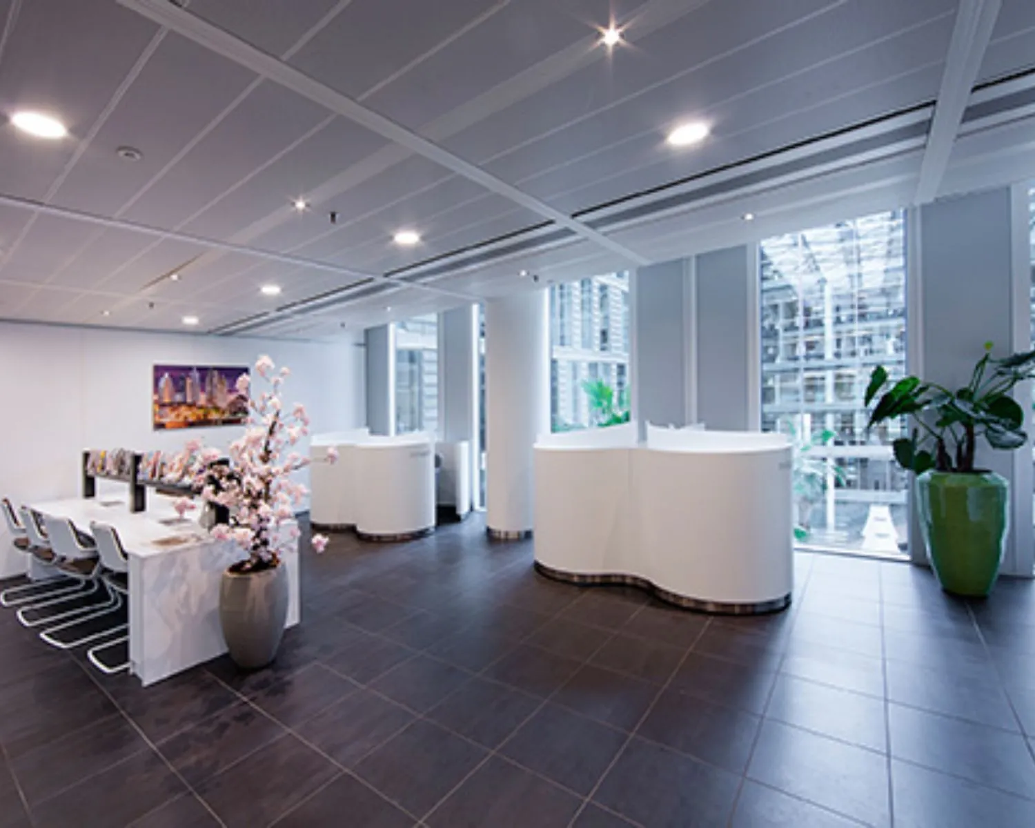 Moderne en lichte ontvangsthal met witte balies, een vergadertafel, en grote kamerplanten bij hoge ramen.
