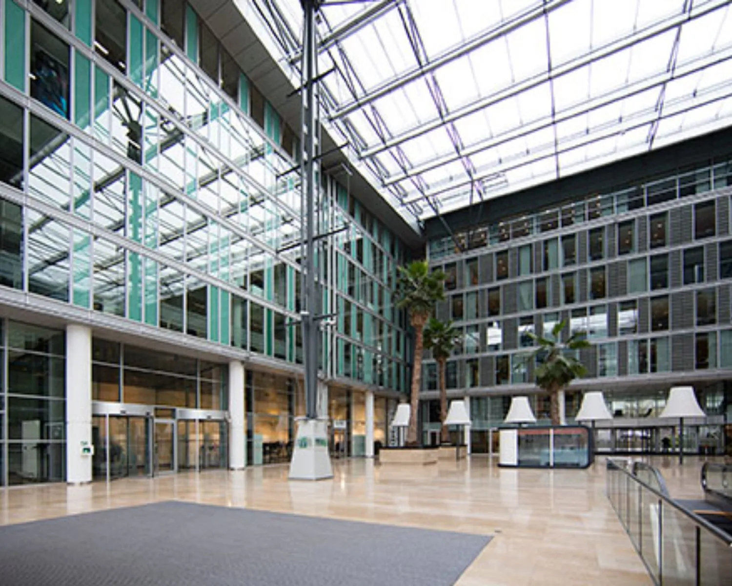 Moderne, glazen atriumhal van een kantoorgebouw met hoge plafonds, palmbomen en veel natuurlijk licht.