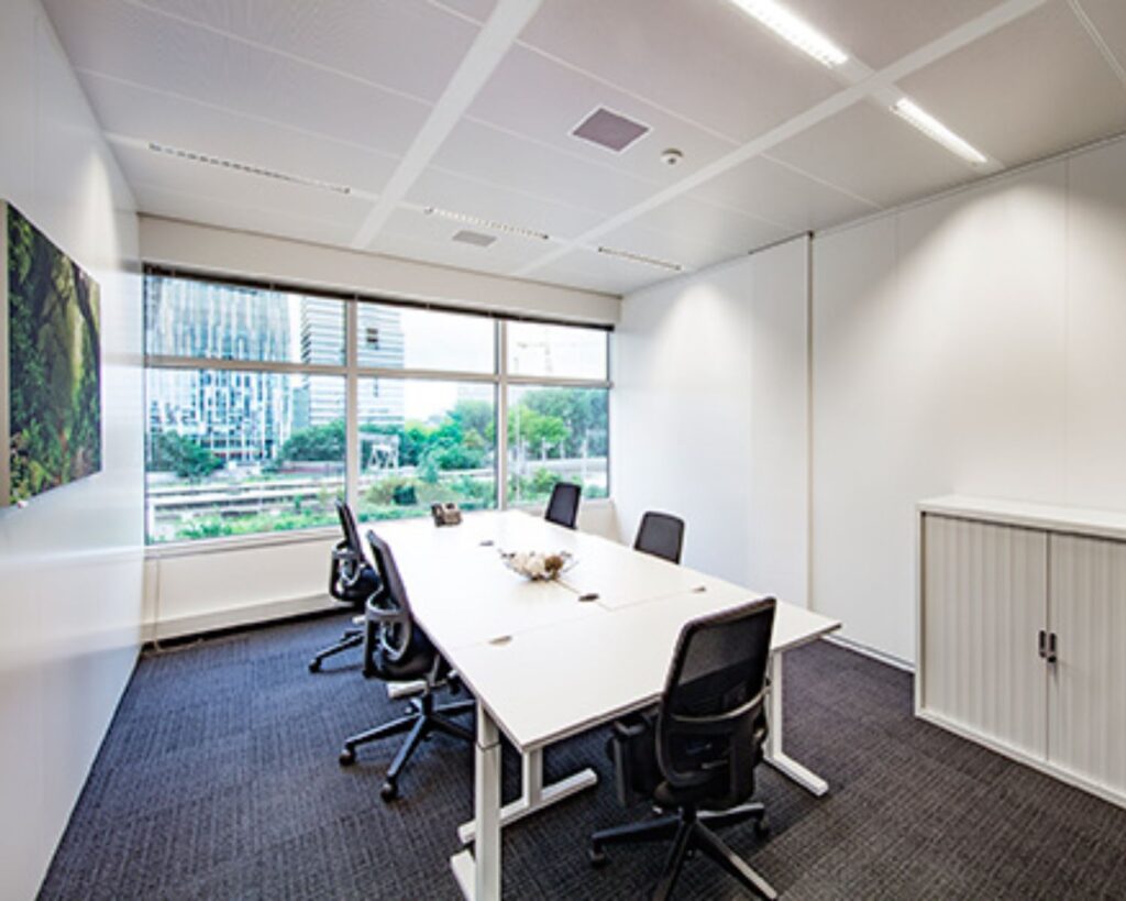 Moderne vergaderruimte met grote ramen, zes stoelen en uitzicht op kantoorgebouwen en groen.