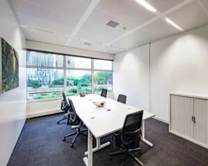 Moderne vergaderruimte met grote ramen, zes stoelen en uitzicht op kantoorgebouwen en groen.