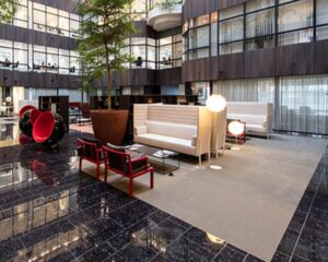 Moderne kantoorlobby met designmeubels, grote planten en glazen kantoorkamers op de achtergrond.